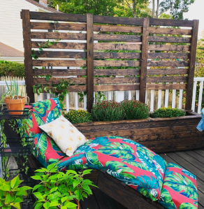 A dark wood planter with a six foot tall trellis on the back. In front of it is a wooden chaise lounge with a brightly colored cushion.